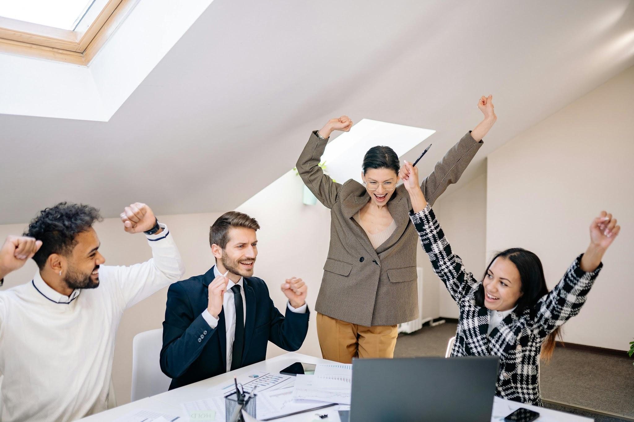 four people sitting and standing round a desk and smiling and cheering