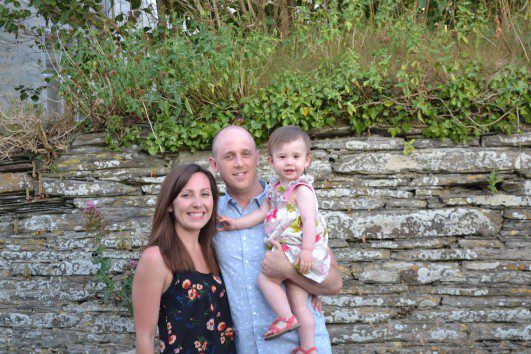 man, woman and toddler stood together in front of wall, smiling at camera