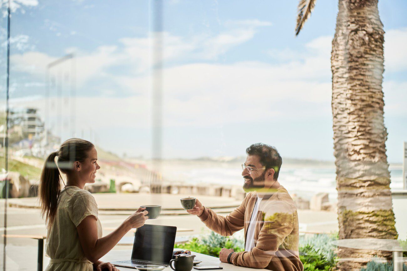 man and woman siting at at table outside cafe, holidng mugs in the air