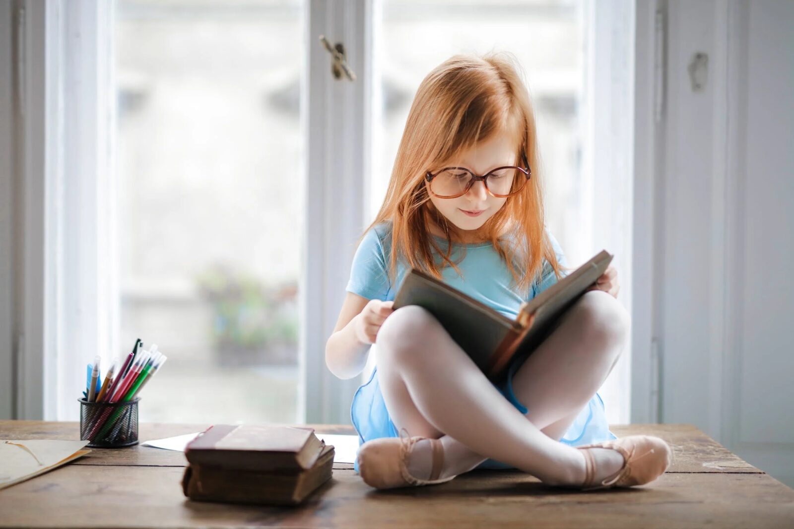 little girl with long red hair and wearing glasses, sitting on a table with a book in her hand