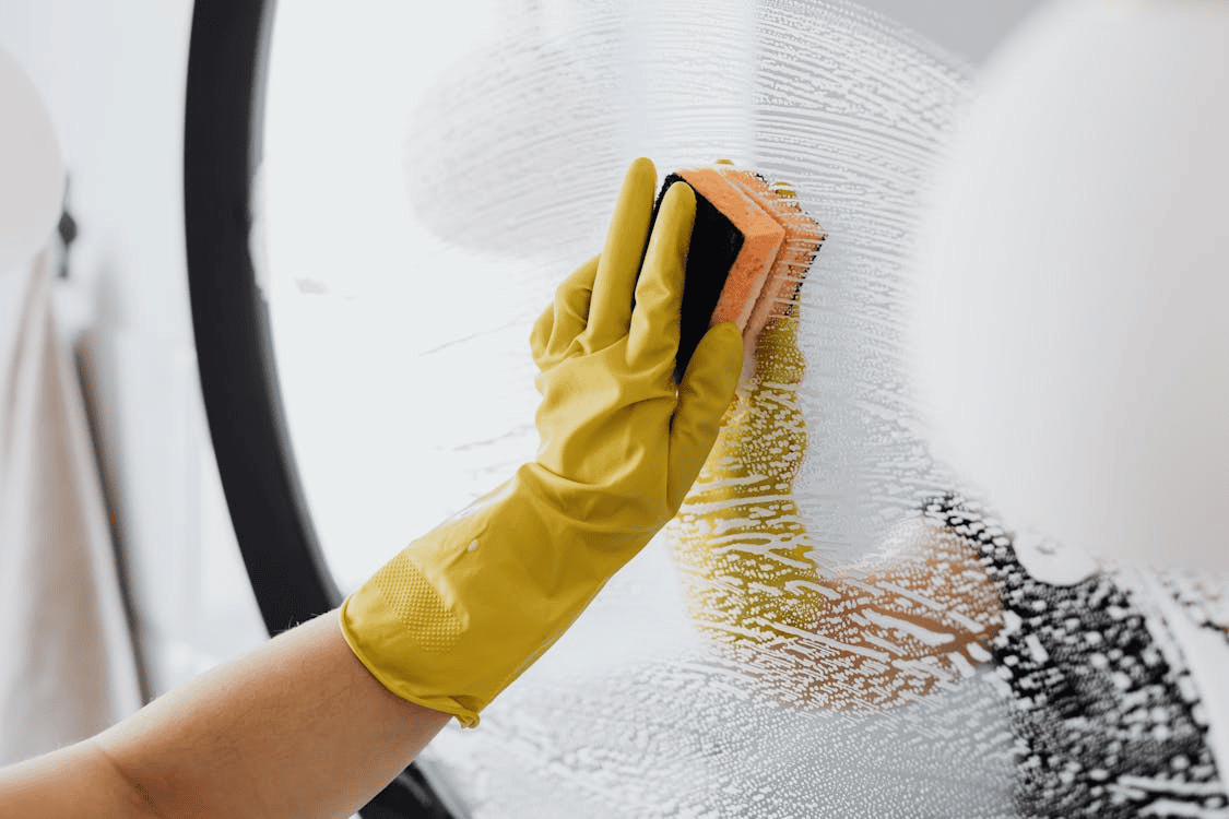person wearing yellow washing up glove cleaning mirror with sponge