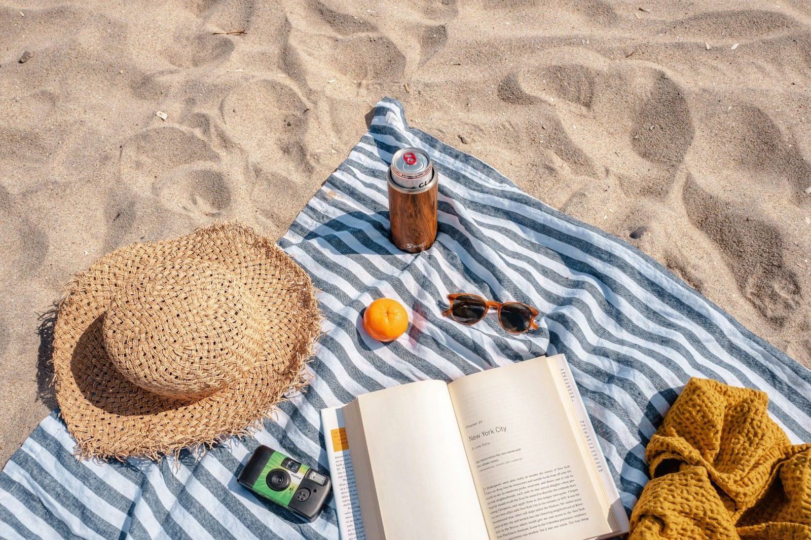 stripey blanket on sand on beach with a hat, book, camera, sunglasses, and drink