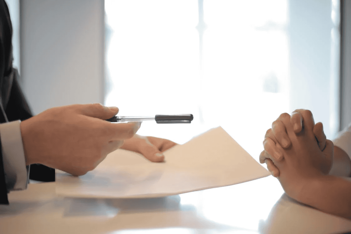 person holding pen and paper, passing them across table to another person
