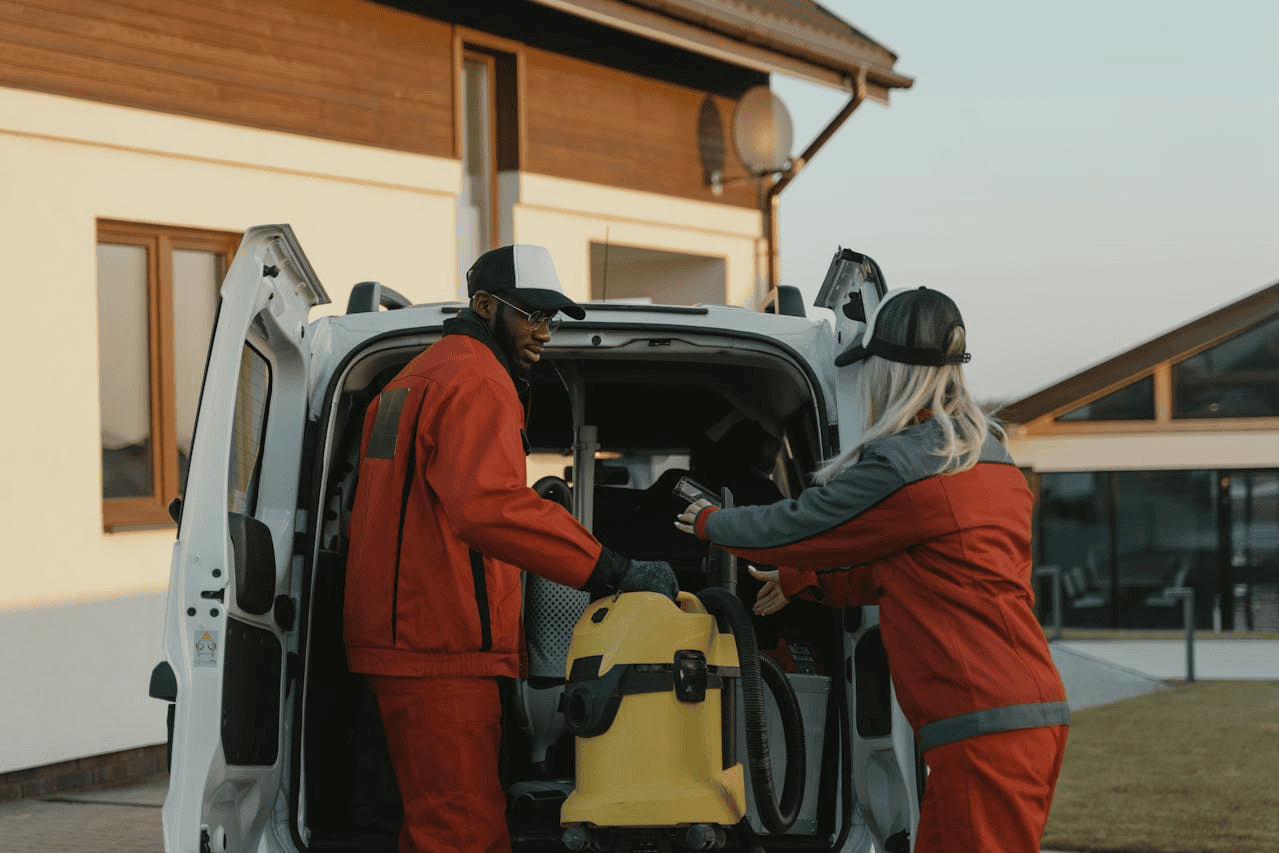 two people in orange and grey unifroms unloading a vacuum cleaner out of a van in front of house