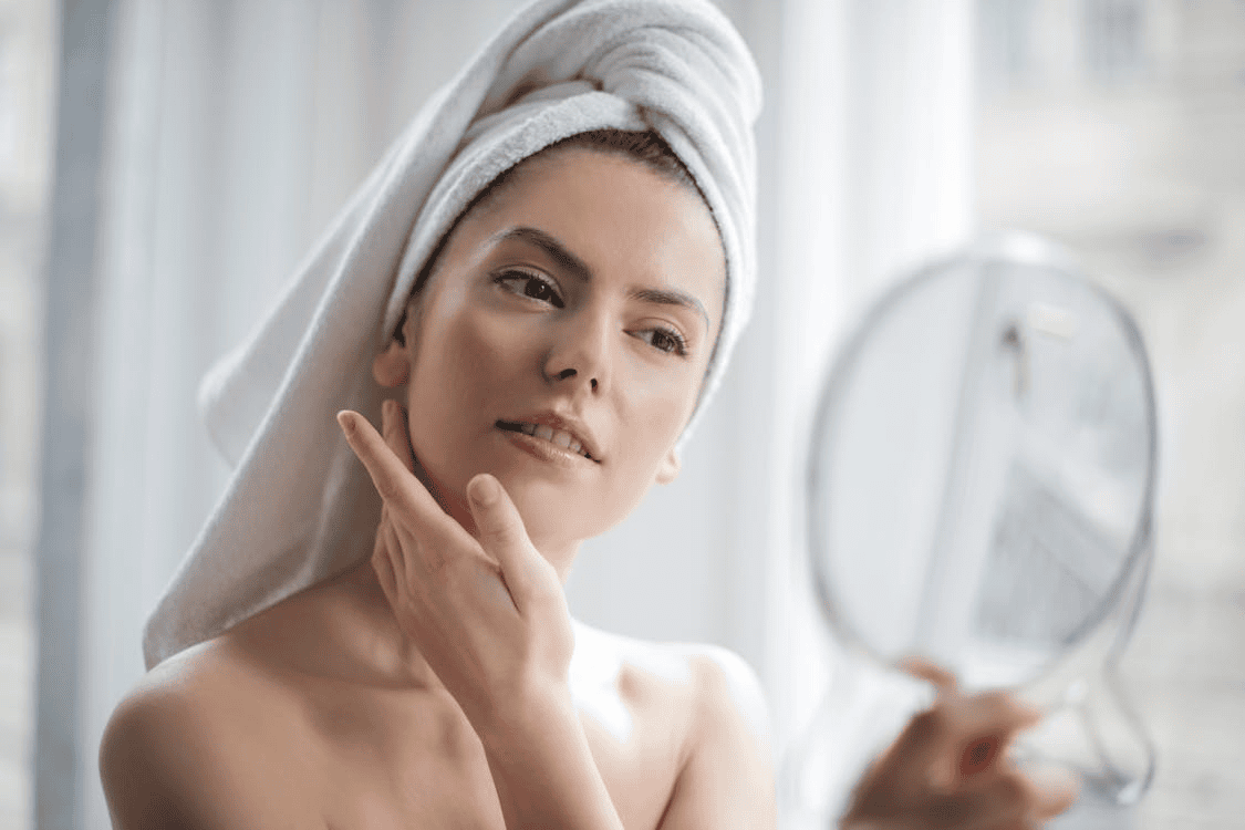 woman with her hair wrapped in a towel, looking in a mirror with her hand touching her face