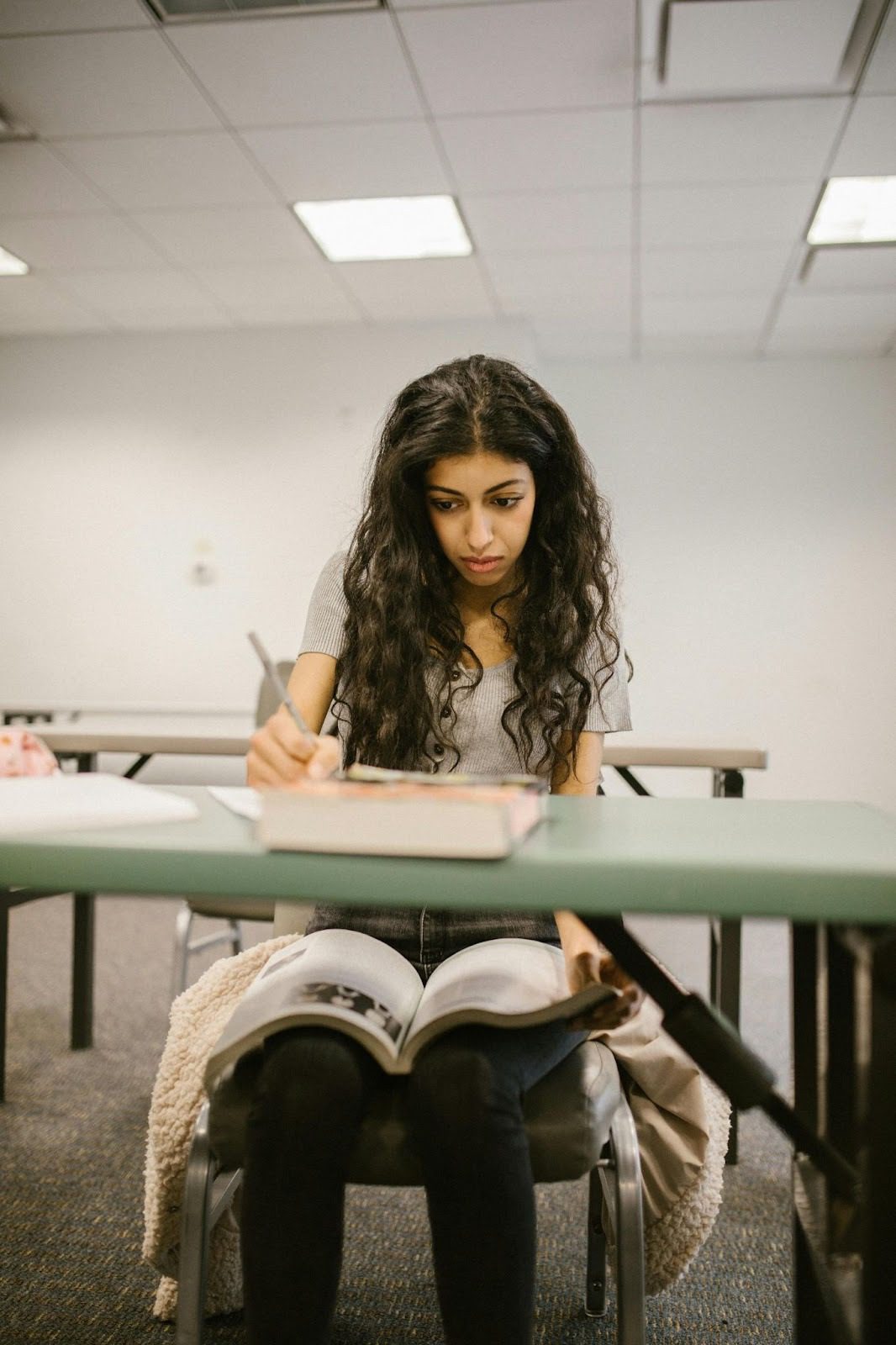 teen girl sitting at desk in classroom writing and with open book on her knee