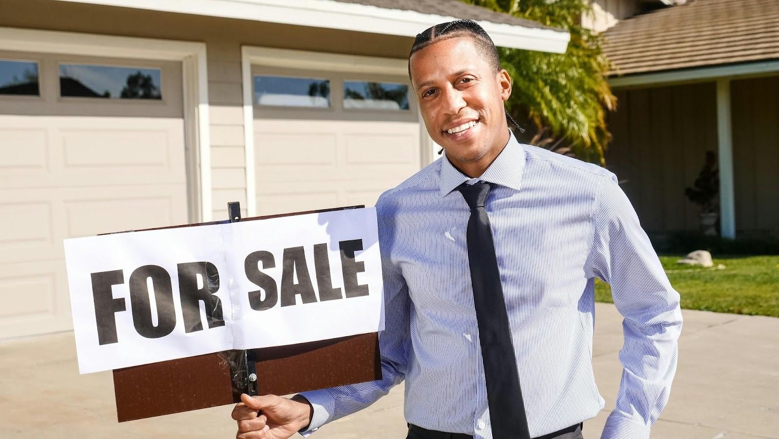 smiling man wearing shirt and tie holding 'for sale' sign outside house