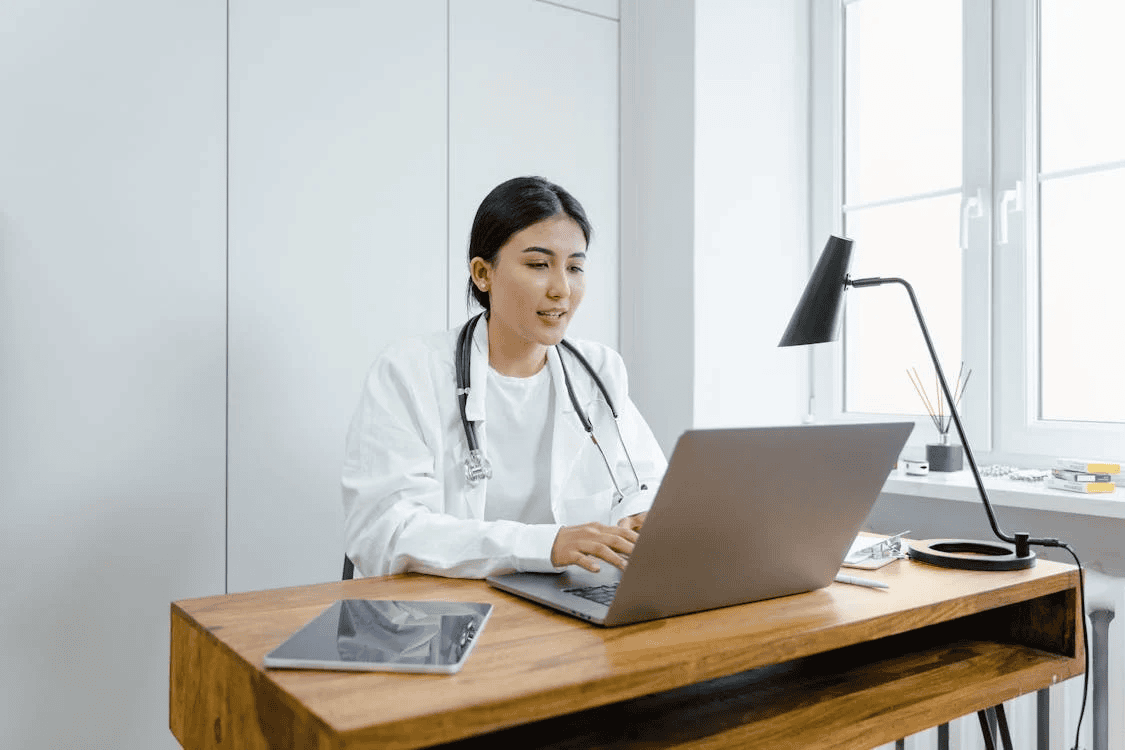 female doctor wearing white coat with stethoscope round neck, sitting at desk and typing on laptop