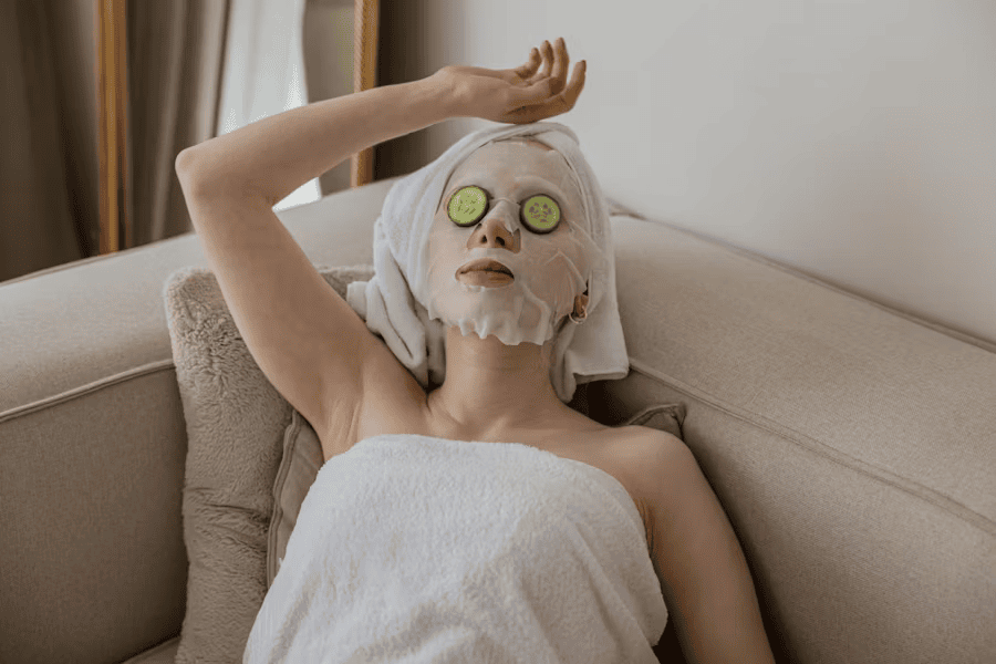 woman lying back on cream sofa, wearing face mask and with cucumber on eyes