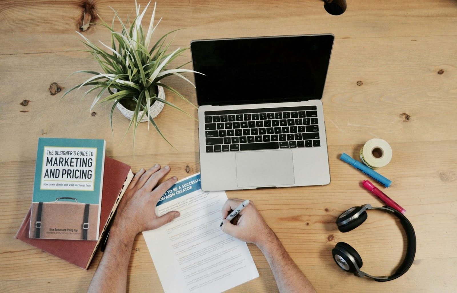 open laptop on table next to marketing book, plant, paper, and person's hands