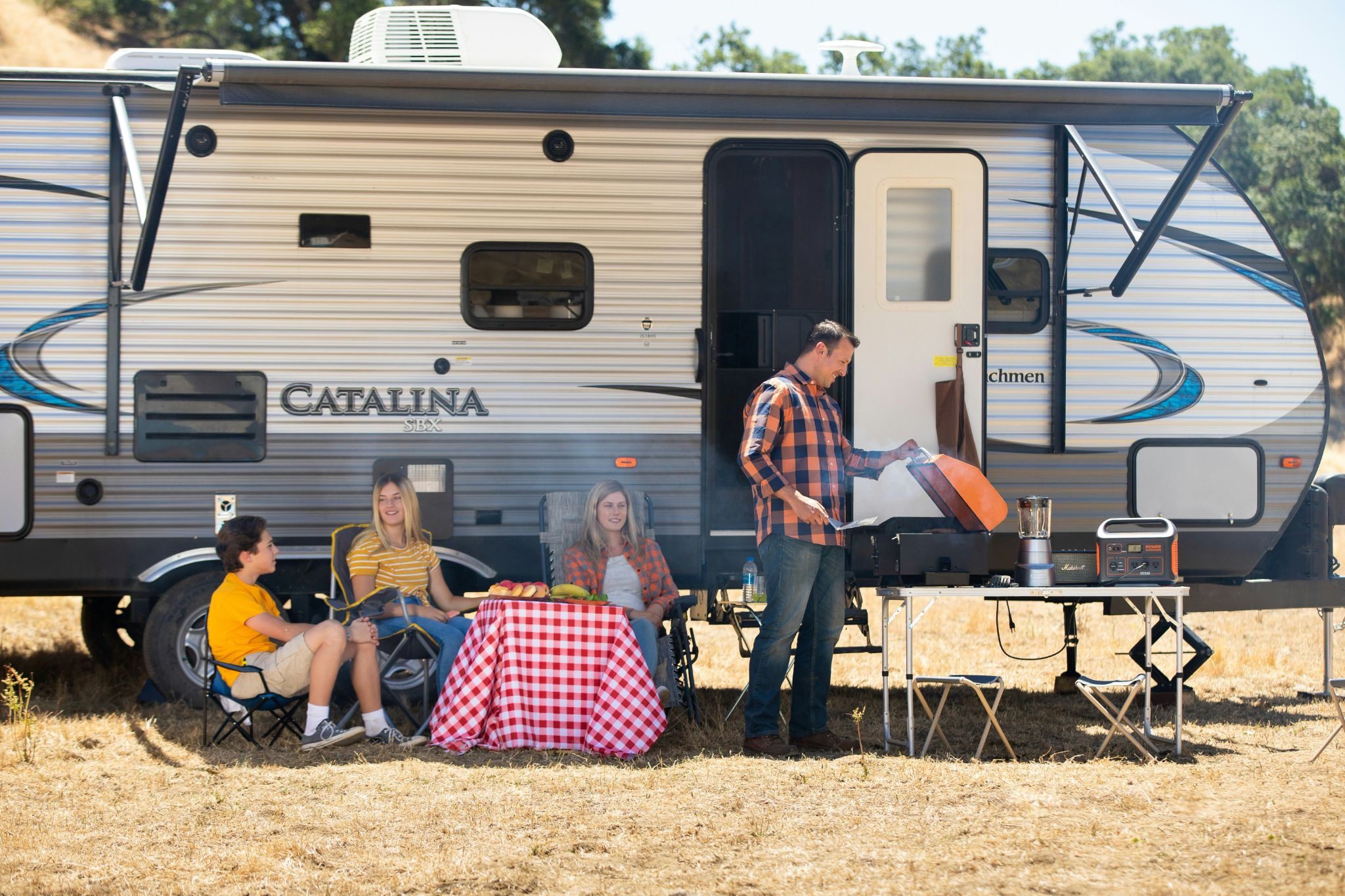 a family sitting next to an RV motorhome with the man cooking on a barbecue