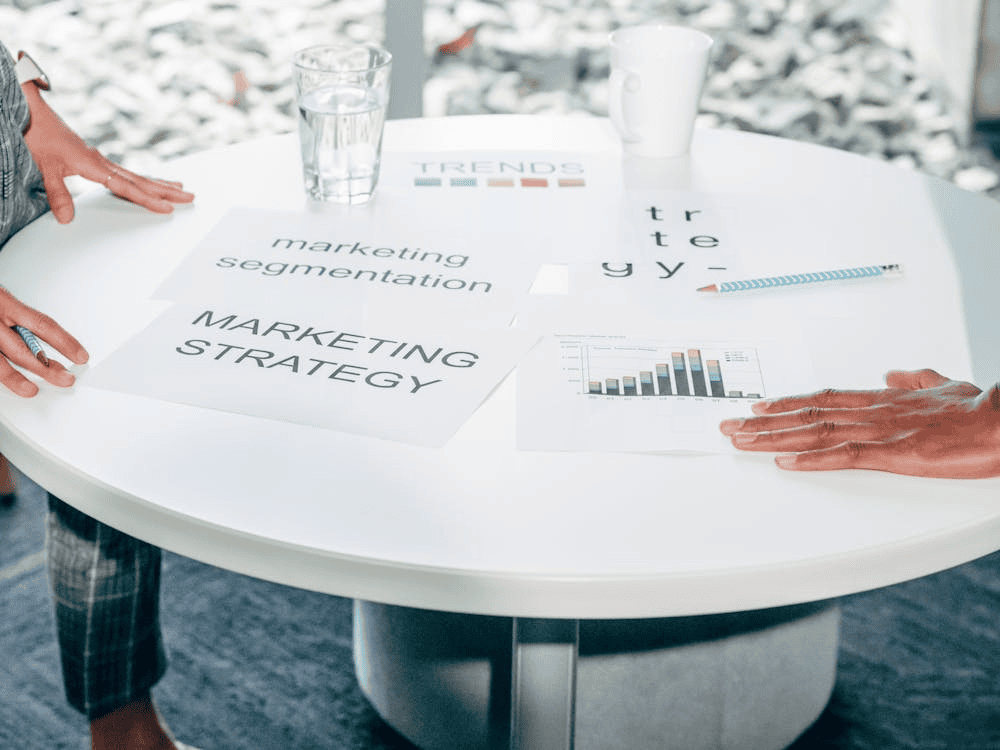 people stood with hands on white table and papers with 'marketing strategy' written on them