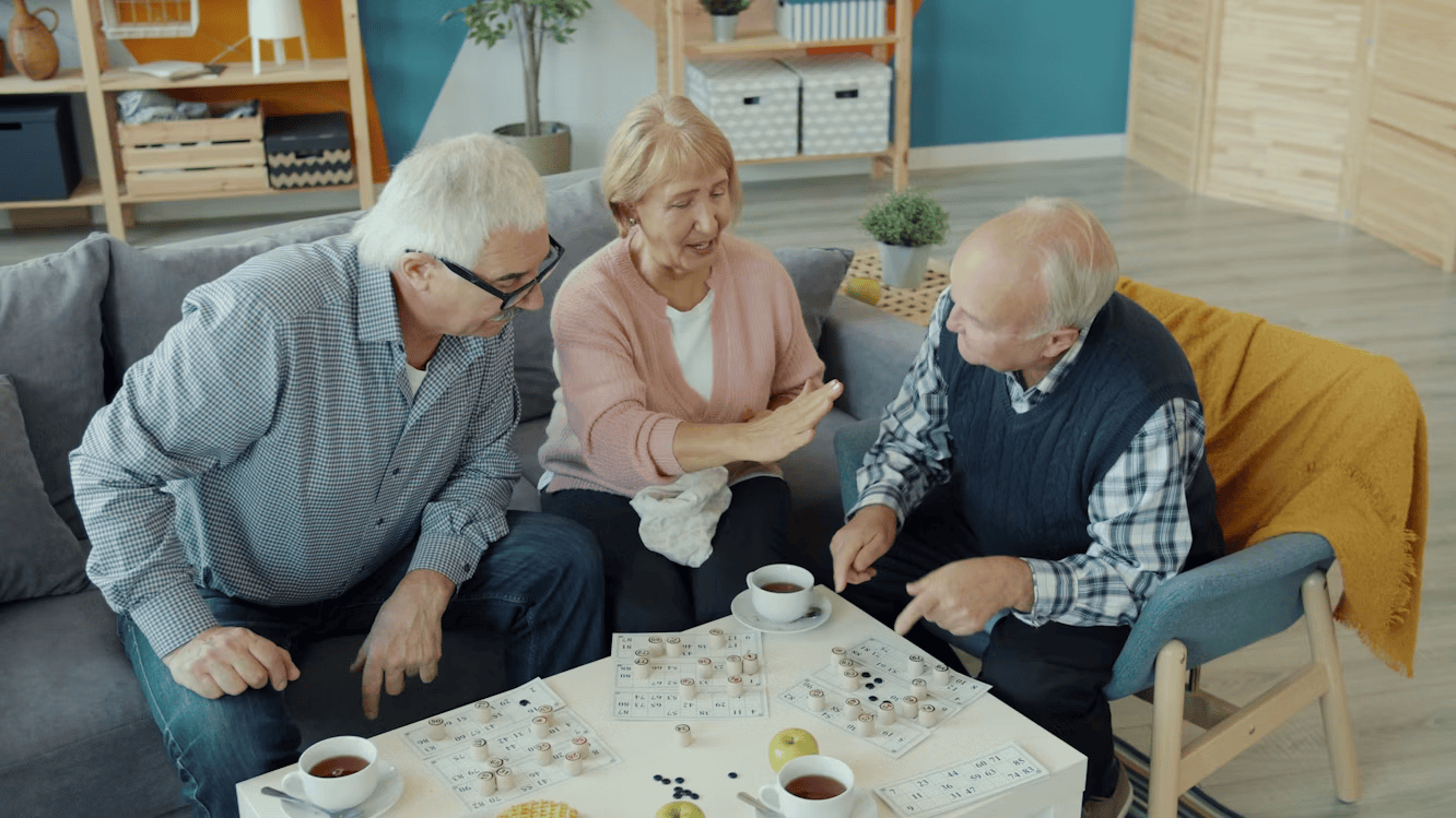 two older men and a woman sitting around a low table, with cups and saucers on and playing a game